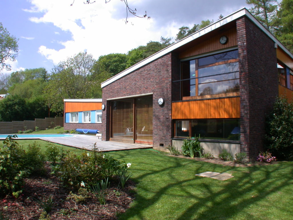 Maison moderne en brique. Son toit est incliné. Elle se trouve dans un jardin avec une terrasse et pisicine.