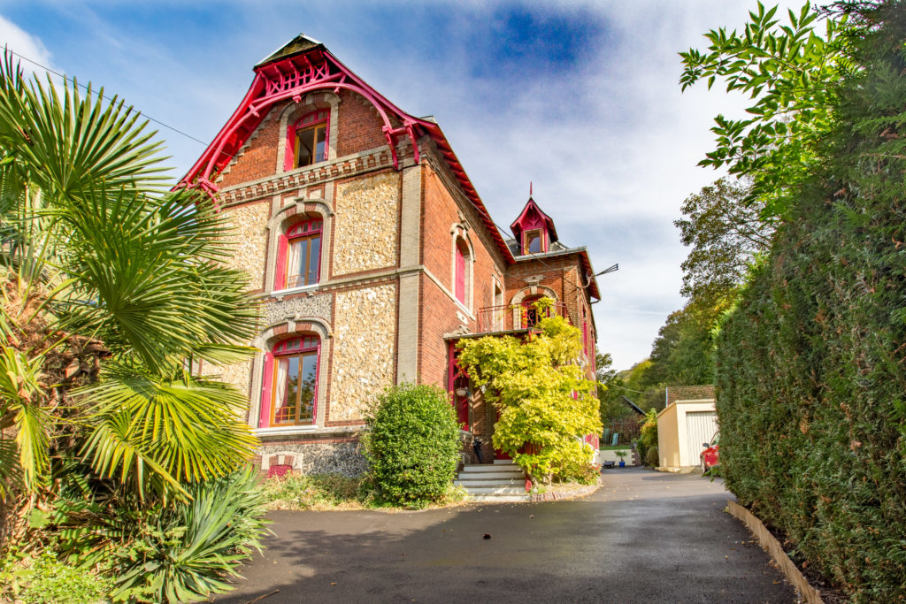 Vue extérieure d'une grande maison en brique de trois étages, avec une toiture et des fenêtres rouges. Elle se trouve dans un jardin.