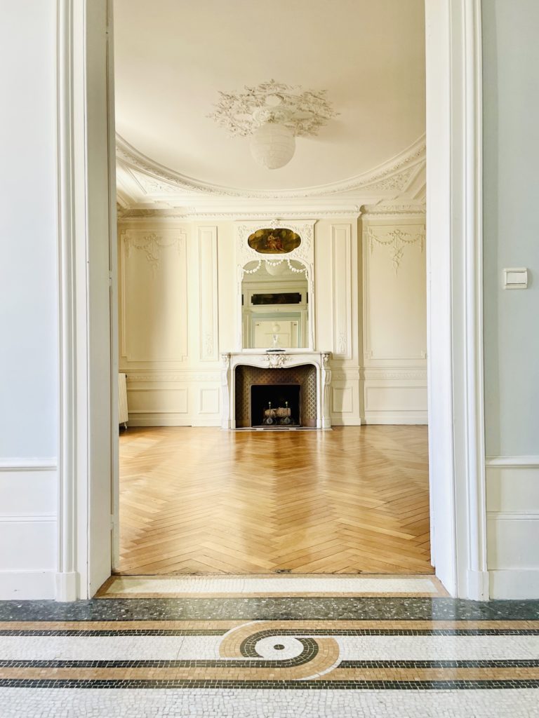 Intérieur de maison, au murs blancs, moulures sur les murs et plafond. Il y a une cheminée. Au sol il y a un parquet et de la mosaïque.