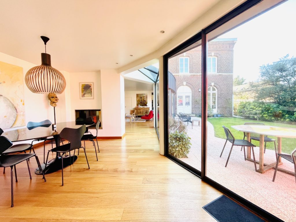 Intérieur d'une maison moderne, donnant sur un salon meublé. Une baie vitrée permet de voir un jardin et une terrasse.