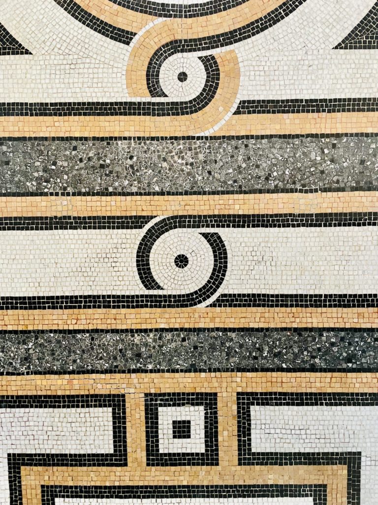 Mosaïque sur le sol d'une maison. Elle représente des formes géométriques. Les motifs sont gris, blancs et noirs.